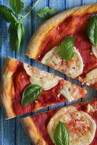 Stock photo showing elevated view of triangular slice of pizza cut from a Margherita pizza topped with a rich tomato sauce, melted circles of buffalo mozzarella and garnished with fresh basil leaves, against a blue tongue and groove effect background.