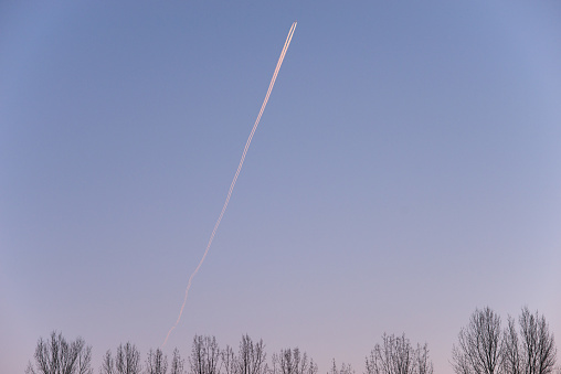 An aircraft and its condensation trail can be seen in a colourful, wintery sky with trees on the ground.