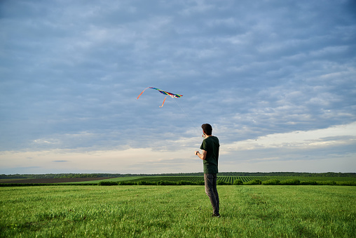 Young brunette skinny man, wearing dark green t-shirt, playing with colorful kite on green field meadow in summer. Kite flying in blue cloudy sky. Family leisure activity