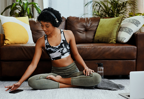 Shot of a young woman wearing headphones and using a cellphone while exercising at home