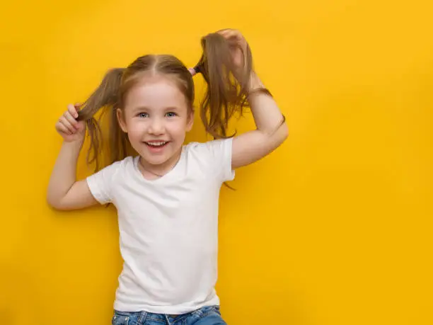 Photo of A naughty little girl in a white T-shirt is holding herself by the hair on a yellow background. Mocap. Copyspace