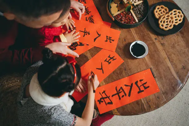 Overhead view of joyful little granddaughter and grandson practising Chinese calligraphy for Chinese New Year on couplets with their grandfather