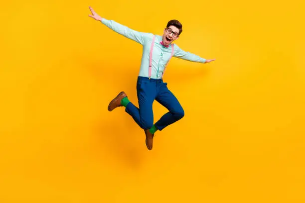 Photo of Full size photo of cool stylish guy jumping high up rejoicing good mood wear specs shirt bow tie suspenders trousers shoes green socks isolated bright yellow color background