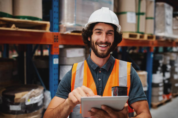 istock Handsome cheerful young caucasian smiling supervisor with white helmet using digital tablet for work while standing in warehouse against goods 1252387789