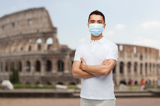 health, safety and pandemic concept - man with crossed arms wearing protective medical mask for protection from virus disease over coliseum in rome, italy background