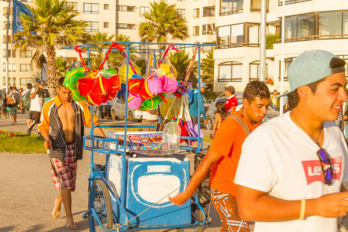 January 14, 2020 - La Serena - Coquimbo, Chile: A young chilean male finishing his working day as Ice Cream Vendor at the beach, in a hot summer day, on back some young latin tourists from Argentinians and Chileans enjoying the summer walking on the seaview pathway in front of the Pacific Ocean.\n\nIn summer, Coquimbo and La Serena beaches are full of Argentinian people from Mendoza and nearest areas, for them, is more easy way access to the ocean, go and drive just 3 hours to Chile for enjoy the summer at the chilean beaches.