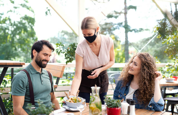 istock Waitress with face mask serving happy couple outdoors on terrace restaurant. 1252395454