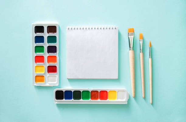 Two sets of color Watercolor paints, three light beige brushes with orange pile and wooden texture and white clear sheet of note book on a light blue background. Horizontal photo. Flat lay minimalistic composition. Back to school, college, education concept. Space for text, copy space. Stationery. Art supplies idea. composición plana de primer plano minimalista horizontal con libro de notas de hoja clara, tres pinceles de color beige claro con pila naranja y textura de madera y dos conjuntos de pintura de acuarela de color sobre un fondo azul claro. objetos en el cen