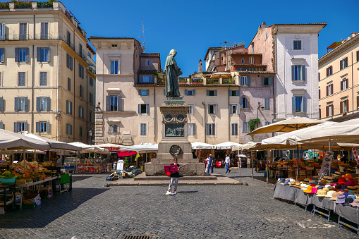 Rome, Italy, June 22 - A street musician plays in the middle of the Campo de Fiori square, among the stalls of the traditional groceries market, under the bronze statue of the religious Giordano Bruno, made by Ettore Ferrari in 1889. In addition to the typical daily and ancient market, the square is also famous for the presence of numerous Italian restaurants, pubs and cafes. Image in High Definition format.