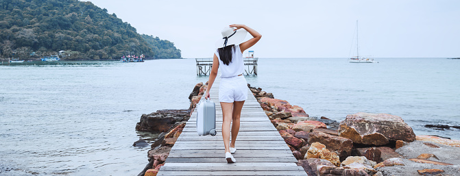 Summer travel vacation concept, Happy traveler asian woman with suitcase bag and straw hat walking on wooden bridge in sea beach at Koh Kood, Trad, Thailand