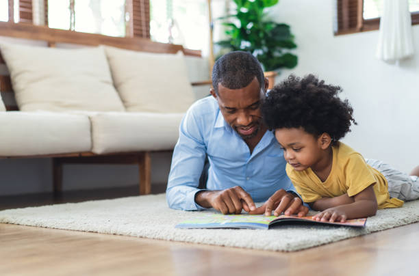 happy african american father and son are reading a book and smiling while lying on floor spending time together at home. children education and development concept. - family reading african descent book imagens e fotografias de stock