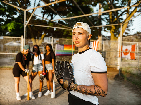 Diverse group of young women practising soft ball at the outdoor city park field. Blonde woman in the foreground, looking at camera.