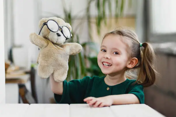 Beautiful little girl is playing with a muppet bear in round shaped glasses and smiling while stay at home.