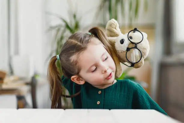 Sweet little girl is playing with a muppet bear in round shaped glasses and smiling while stay at home.
