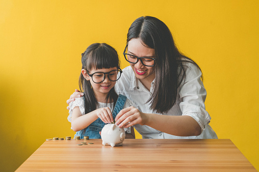 Image of Little Asian Cute Girl Putting Money To Piggy bank Sitting With Mother in living room.