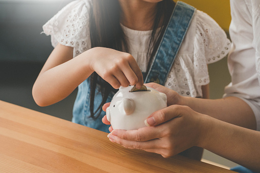 Image of Little Asian Cute Girl Putting Money To Piggy bank Sitting With Mother in living room.