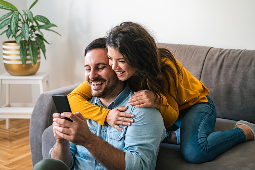 Smiling girl embracing from behind her happy boyfriend while using mobile phone and laughing together at home