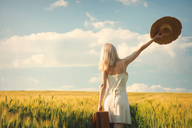 istock Blode woman in hat with suitcase in wheat field in sunset time 1252475476
