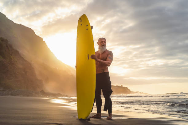 happy fit senior zabawy surfing w czasie zachodu słońca - sporty brodaty człowiek szkolenia z deski surfingowej na plaży - aktywnych osób starszych styl życia i ekstremalnych koncepcji sportu - surfing surf wave men zdjęcia i obrazy z banku zdjęć
