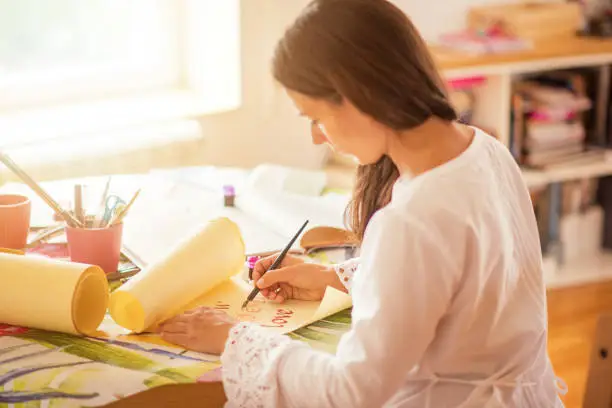 Beautiful young woman sitting at table in her home and enjoying writing calligraphy. Young artist relaxing at home and enjoying calligraphy.