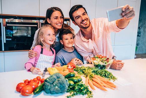 Happy young family of four cooking together and making a selfie in modern kitchen.