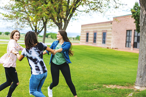 In Western Colorado Group of Generation Z Multi-Ethnic Female Friends Running in Circles Holding Hands on Green Grassy Area in School Yard Near High School Building Part of a Series (Shot with Canon 5DS 50.6mp photos professionally retouched - Lightroom / Photoshop - original size 5792 x 8688 downsampled as needed for clarity and select focus used for dramatic effect)