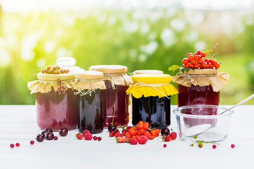 Fruit and berry preserves in the glass jars and  raw strawberries , cherries , rowans and red currants berries on a white wooden table, on the nature background, organic meal and dessert concept, natural sunlight