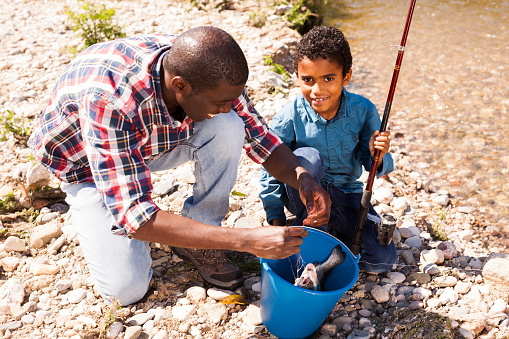 Positive African man and little boy standing near river and holding fishing rod and bucket of catch