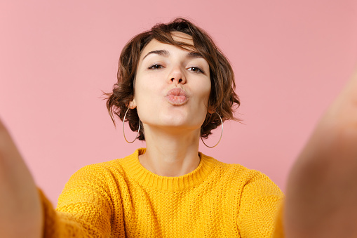 Close up of pretty young brunette woman in yellow sweater posing isolated on pink background. People lifestyle concept. Mock up copy space. Doing selfie shot on mobile phone, blowing sending air kiss