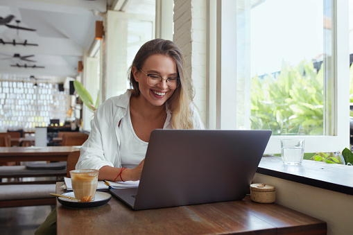 Woman. Online Work At Cafe. Happy Girl In Casual Clothes And Glasses With Laptop Looking At Screen. Modern Digital Technologies For Remote Job, Education And Comfortable Lifestyle In City.