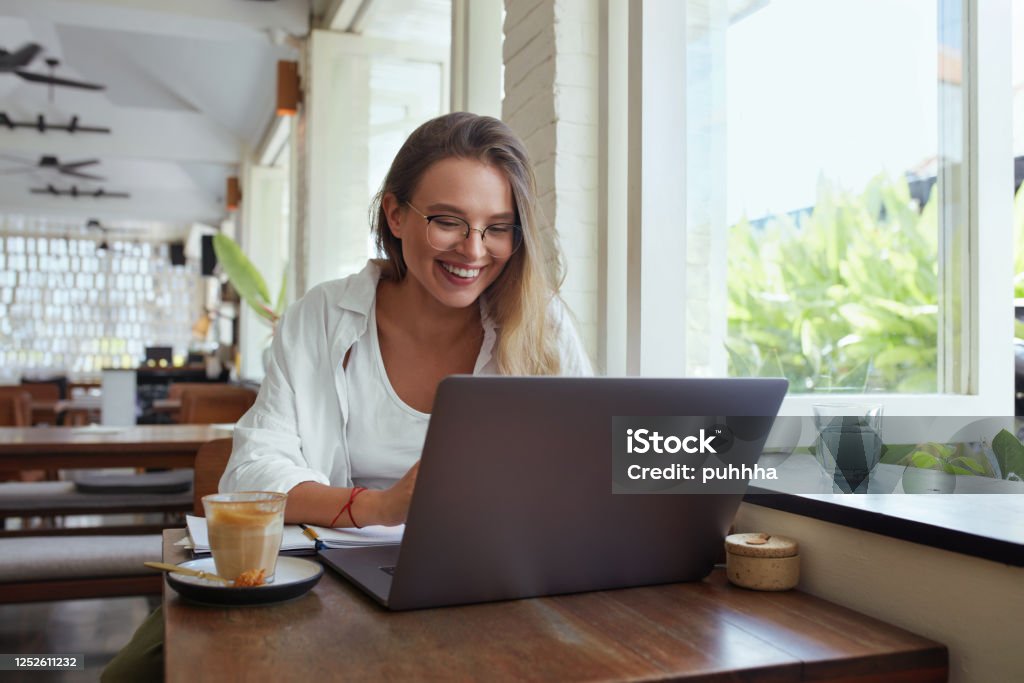Woman. Online Work At Cafe. Happy Girl In Casual Clothes And Glasses With Laptop Looking At Screen. Modern Digital Technologies For Remote Job, Education And Comfortable Lifestyle In City. One Person Stock Photo