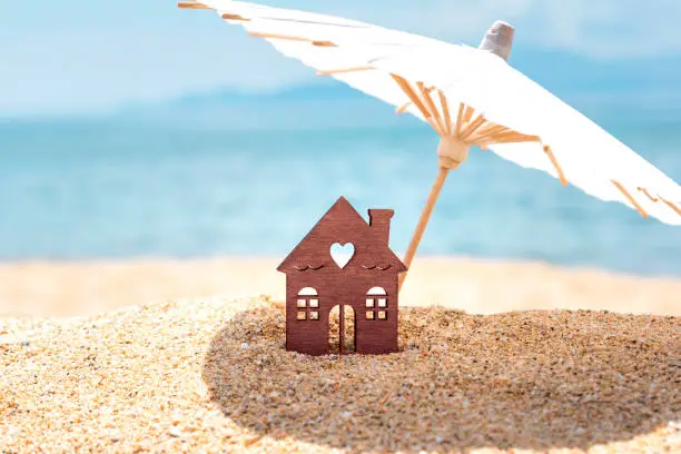 Photo of Miniature house and umbrella on beach, blue sea and sky on blurred background. Real estate, sale or property investment concept. Symbol of dream home for family. Copy space