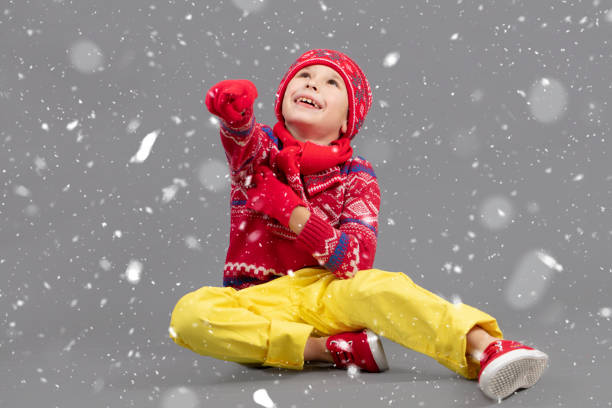 Cheerful Child in warm clothes on a winter background. A boy in a red knitted hat and sweater. Fashionable boy in the studio in white snowflakes