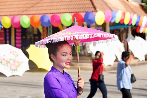 Smiling young thai woman with pink paper umbrella in traditional fashion captured in street in Chiang Mai. Woman is sitting on a bicycle out of frame and is part of a row of women. Scne is at annual umbrella an dart festival. In background tourists are walking.