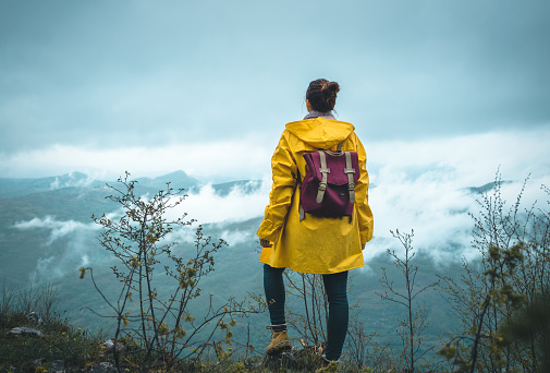 Woman hiking in nature in yellow raincoat standing on mountain peak looking to view