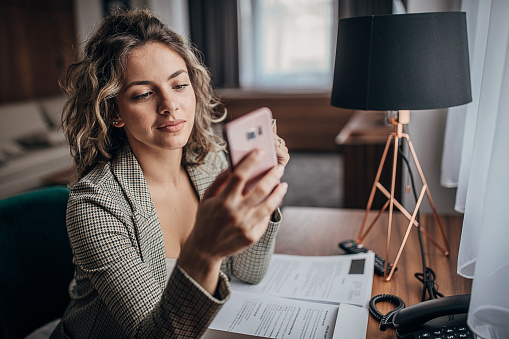 One young beautiful businesswoman working from hotel room on business trip, using smart phone.