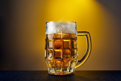 Cold craft light Beer in a mug with drops on a dark table. Pint of Beer on yellow color background. Close up.