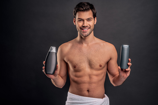 Handsome young man isolated. Portrait of shirtless muscular man is standing on grey background with shampoo in hands. Men care concept