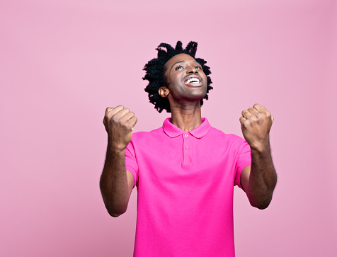Summer portrait of surprised afro american young man wearing pink polo shirt, looking up with raised fists. Studio shot on pink background.