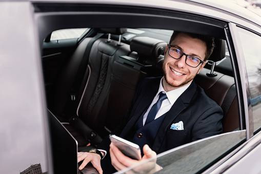 Smiling bearded businessman wearing suit and glasses sitting in back seat of car, looking out of window, holding smart phone