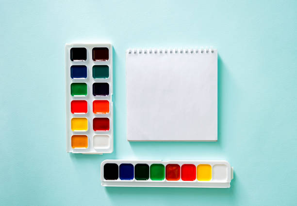 Two sets of color Watercolor paints and white clear sheet of note book on a light blue background. Horizontal photo. Flat lay minimalistic composition. Back to school, college, education concept. Space for text, copy space. Stationery. Art supplies idea. composición plana de primer plano minimalista horizontal con libro de notas de hoja clara y dos conjuntos de pintura de acuarela de color sobre un fondo azul claro. objetos en el centr de la foto. papelería. foto horizontal. regreso a la escuela, a la un