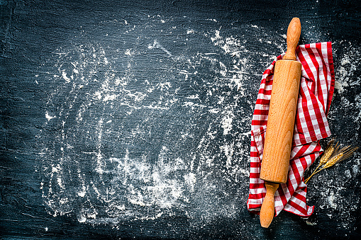 Cooking or kitchen backgrounds: wooden rolling pin on checkered napkin shot from above on black background with spilled white flour. The composition is at the right of an horizontal frame leaving useful copy space for text and /or logo. High resolution 42Mp studio digital capture taken with SONY A7rII and Zeiss Batis 40mm F2.0 CF lens