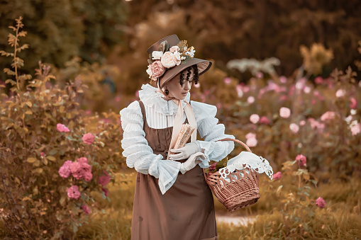 Outdoor close up portrait of beautiful woman wearing wide brim ribbon tie straw hat, vintage dress. Model posing in the blooming rose garden. Woman wearing retro dress with basket and fan.