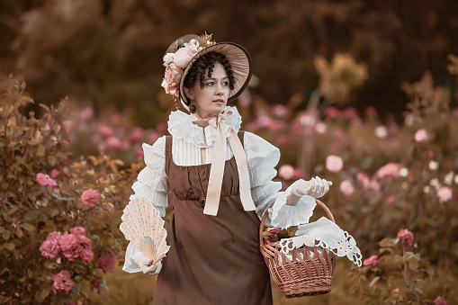 Outdoor close up portrait of beautiful woman wearing wide brim ribbon tie straw hat, vintage dress. Model posing in the blooming rose garden. Woman wearing retro dress with basket and fan.