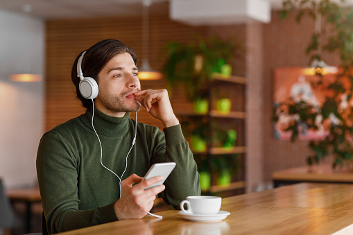 Pensive young man in headphones listening to music, using mobile phone, drinking coffee, cafe interior, empty space