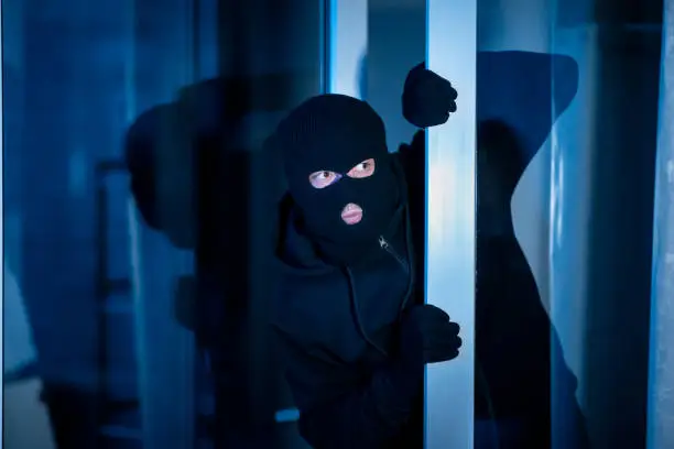 Criminal Concept. Masked burglar peeking into the house through an open glass door or window, copy space