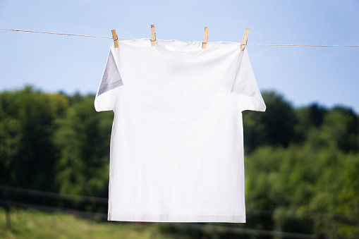 White t-shirt mock-up hanging on the rope with clothespins on summer background. Mockup ready to place your design in front of blue sky and green trees