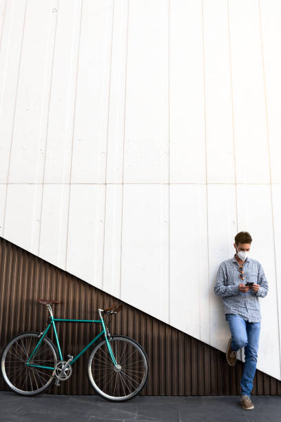 Attractive man with mask and fixed gear bicycle standing on a two-color modern wall with his mobile phone. stock photo