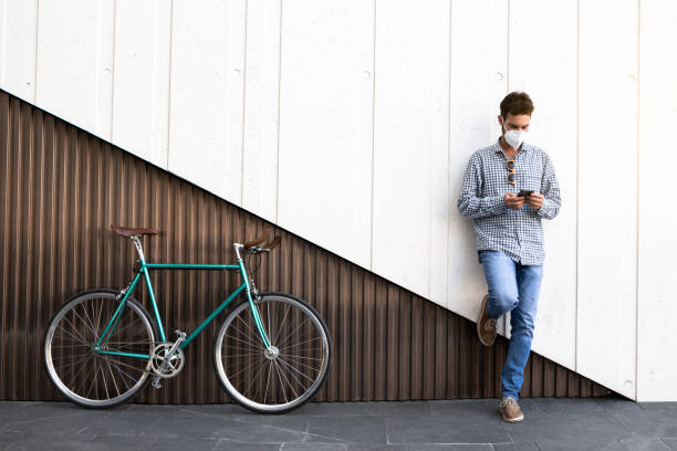 Attractive man with mask and fixed gear bicycle standing on a two-color modern wall with his cell phone. stock photo