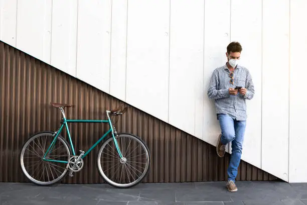 Attractive man with mask and fixed gear bicycle standing on a two-color modern wall with his cell phone. Coronavirus period. Horizontal photography, hipster style.